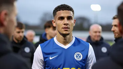 A Barrow AFC player in a blue kit with white sleeves, standing in Holker Street's Fan Zone before the match, surrounded...