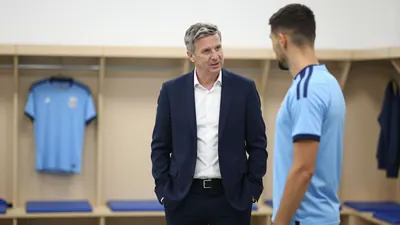 A Barrow manager in a navy blue suit and white shirt, mid-conversation with a player in a light blue training kit, both...