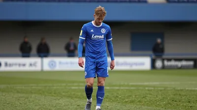 A Barrow AFC player in a blue jersey with a Newfoundland and Labrador logo on the front, looking disappointed as he walks...
