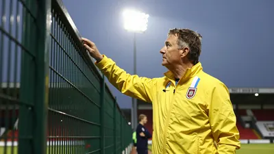 A 35-year-old stadium manager, wearing a bright yellow jacket and a Barrow AFC kit with blue and white stripes, stands in...