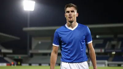 A close-up of a Barrow player's face, with a determined expression, wearing a blue jersey with a white collar and a white...