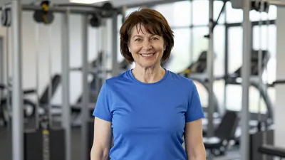 A middle-aged woman with short brown hair, wearing a solid blue Barrow AFC training top, smiles warmly while standing in...