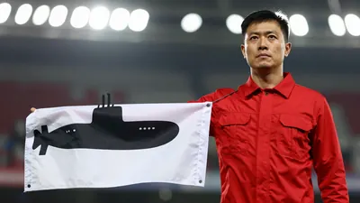 A mid-30s male Barrow staff member, waist-up, wearing a solid red kit, looking determined as he holds a folded flag with...
