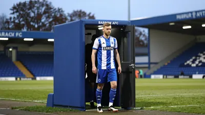 A photograph of a Barrow AFC coach arriving at the stadium, with a team player standing at the door, wearing the team's...