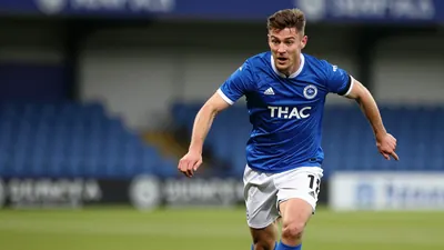 A tight action shot of a young, muscular defender in a Barrow AFC kit (solid blue shirt, white shorts) sprinting down the...