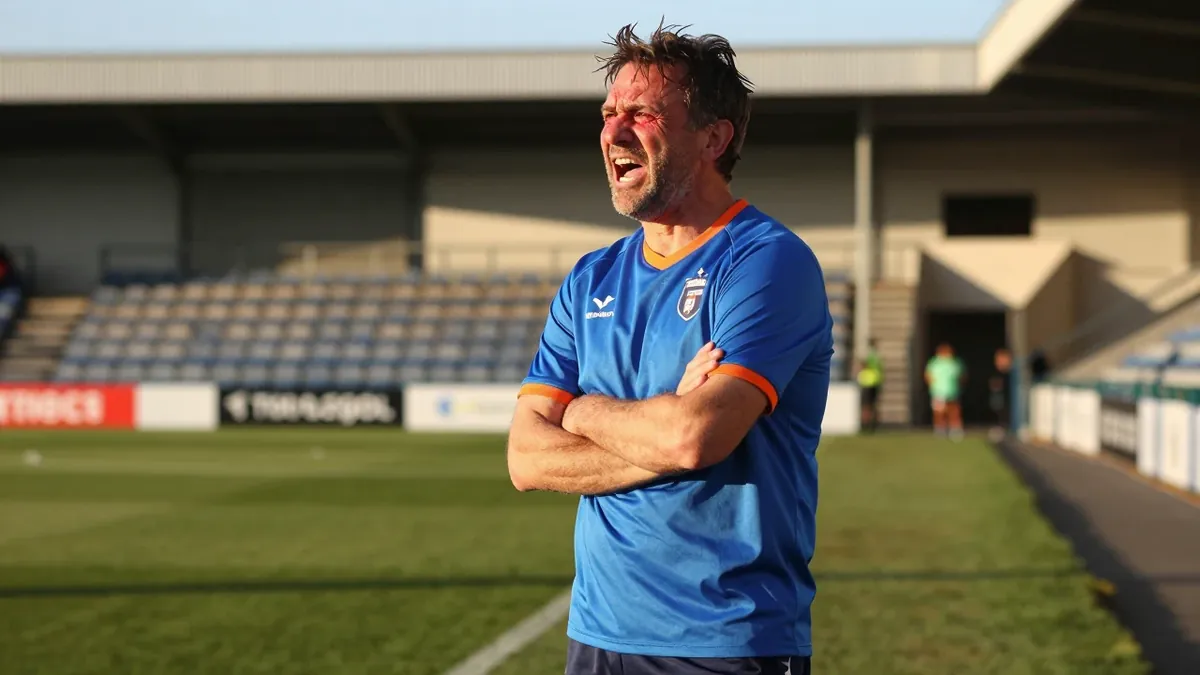 A 45-year-old, ruggedly-built, unshaven Barrow assistant coach, dressed in a plain blue cotton training top with orange...