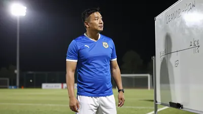 A 35-year-old male coach with a strong build, wearing a blue jersey and white shorts, standing in the middle of the...