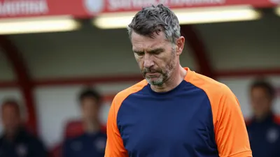 A rugged, 40-year-old Barrow AFC manager, in a plain blue and orange training top, stands pitchside, eyes cast downward...