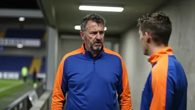A 50-year-old, ruggedly-built, Barrow AFC assistant coach, with a stern expression, stands in the dimly lit, tunnel-like...