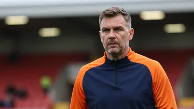 A determined, mid-30s Barrow AFC manager with a strong build, wearing a plain blue and orange training top, stands...