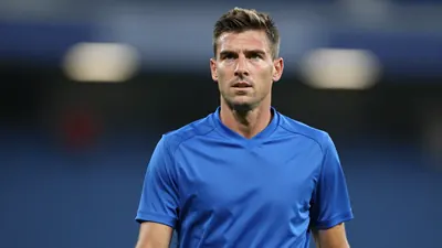 A mid-30s male Barrow player in a solid blue shirt, waist-up, looking determined, stadium lights blurred behind him...