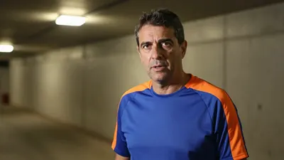 A 50-year-old male head coach, dressed in a plain unmarked blue and orange polyester training top, stands in the low-lit...
