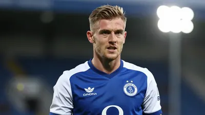 A 25-year-old Barrow AFC player, wearing a blue and white kit, in a waist-up shot, with a determined expression, taken...