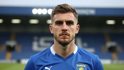 A head-and-shoulders photo of a young male coach, 30-40 years old, wearing Barrow AFC’s blue kit, taken at a stadium...