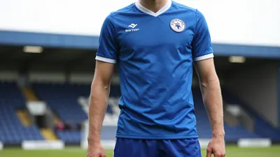 A close-up shot of a Barrow AFC player in the East Stand, wearing a blue jersey with a white collar, and a pair of blue...